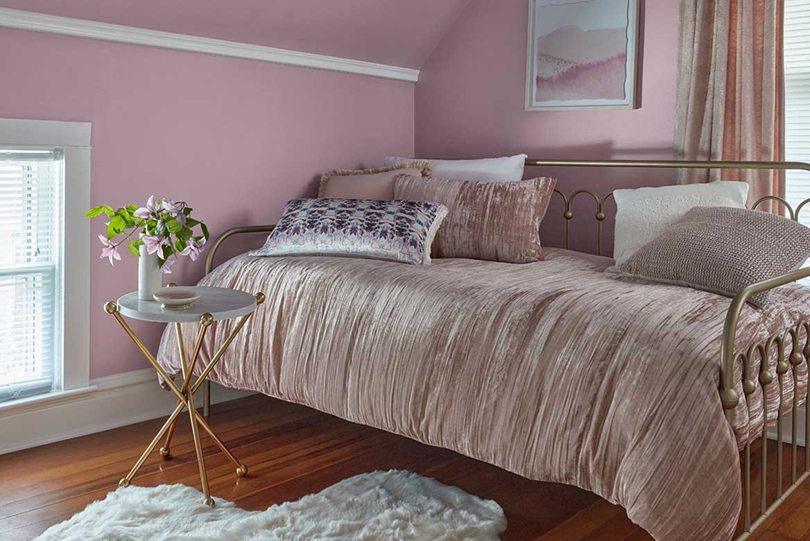 Attic bedroom with pastel lavender walls, daybed covered in velvety soft blanket. A small round table.